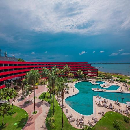 Royal Tulip Brasilia Alvorada Hotel Luaran gambar The photo shows a modern hotel building with a striking red exterior, surrounded by lush greenery. In the foreground, there is a large, creatively shaped swimming pool, with lounge chairs arranged around it. The backdrop features a body of water, pos