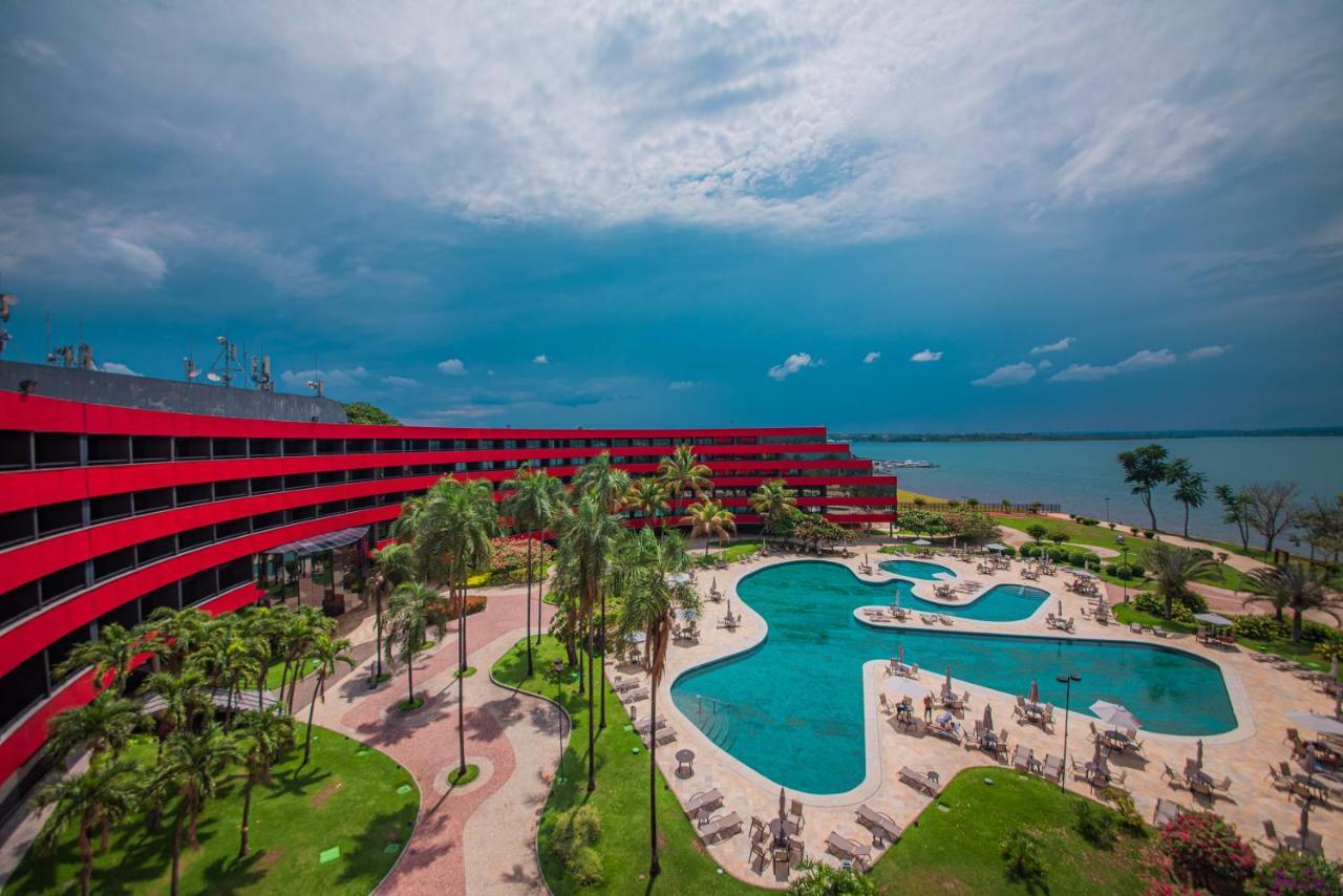 Royal Tulip Brasilia Alvorada Hotel Luaran gambar The photo shows a modern hotel building with a striking red exterior, surrounded by lush greenery. In the foreground, there is a large, creatively shaped swimming pool, with lounge chairs arranged around it. The backdrop features a body of water, pos