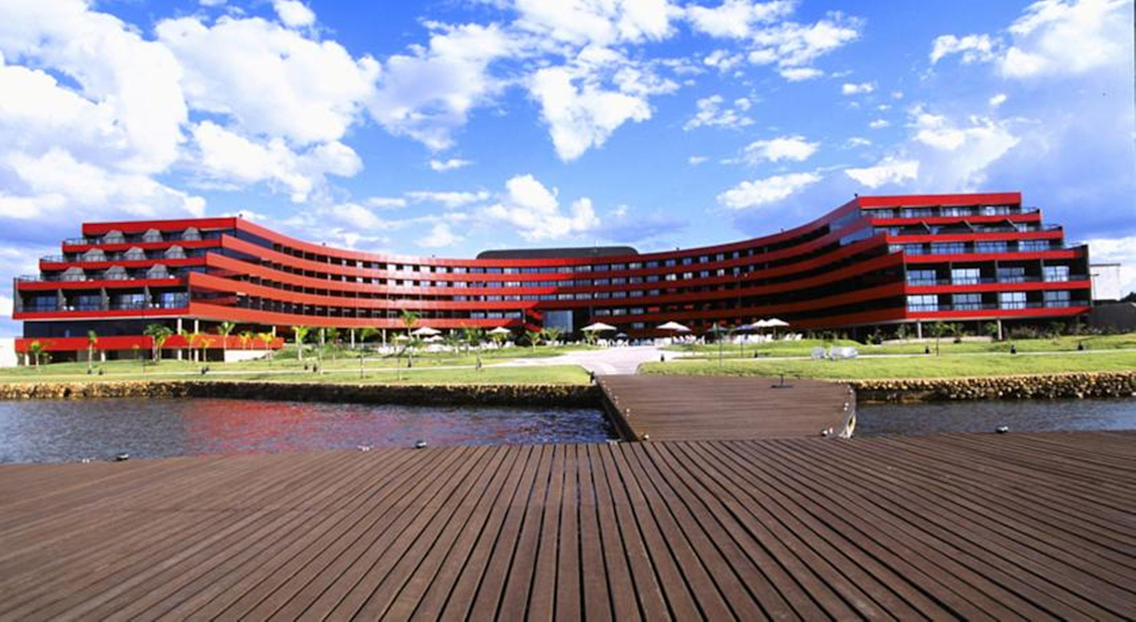 Royal Tulip Brasilia Alvorada Hotel Luaran gambar The image shows a modern, curved building with a striking red facade, situated near a body of water. The architecture features multiple levels with large windows that create a sleek and contemporary appearance. In front of the building, there is a wo
