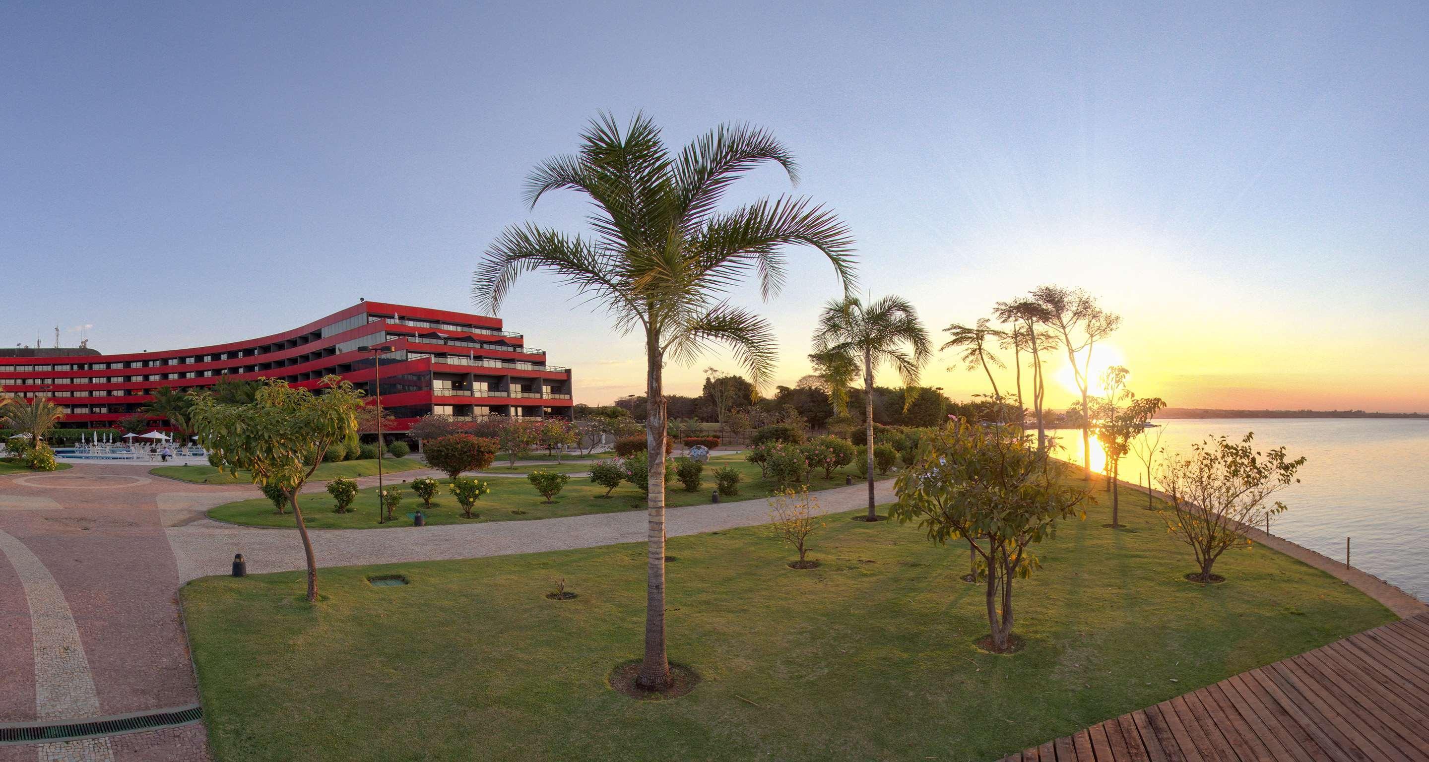 Royal Tulip Brasilia Alvorada Hotel Luaran gambar The photo depicts a scenic view featuring a red multi-story building alongside a serene waterfront. In the foreground, there are several palm trees and lush green lawns, creating a tropical atmosphere. The sun is setting in the background, casting wa