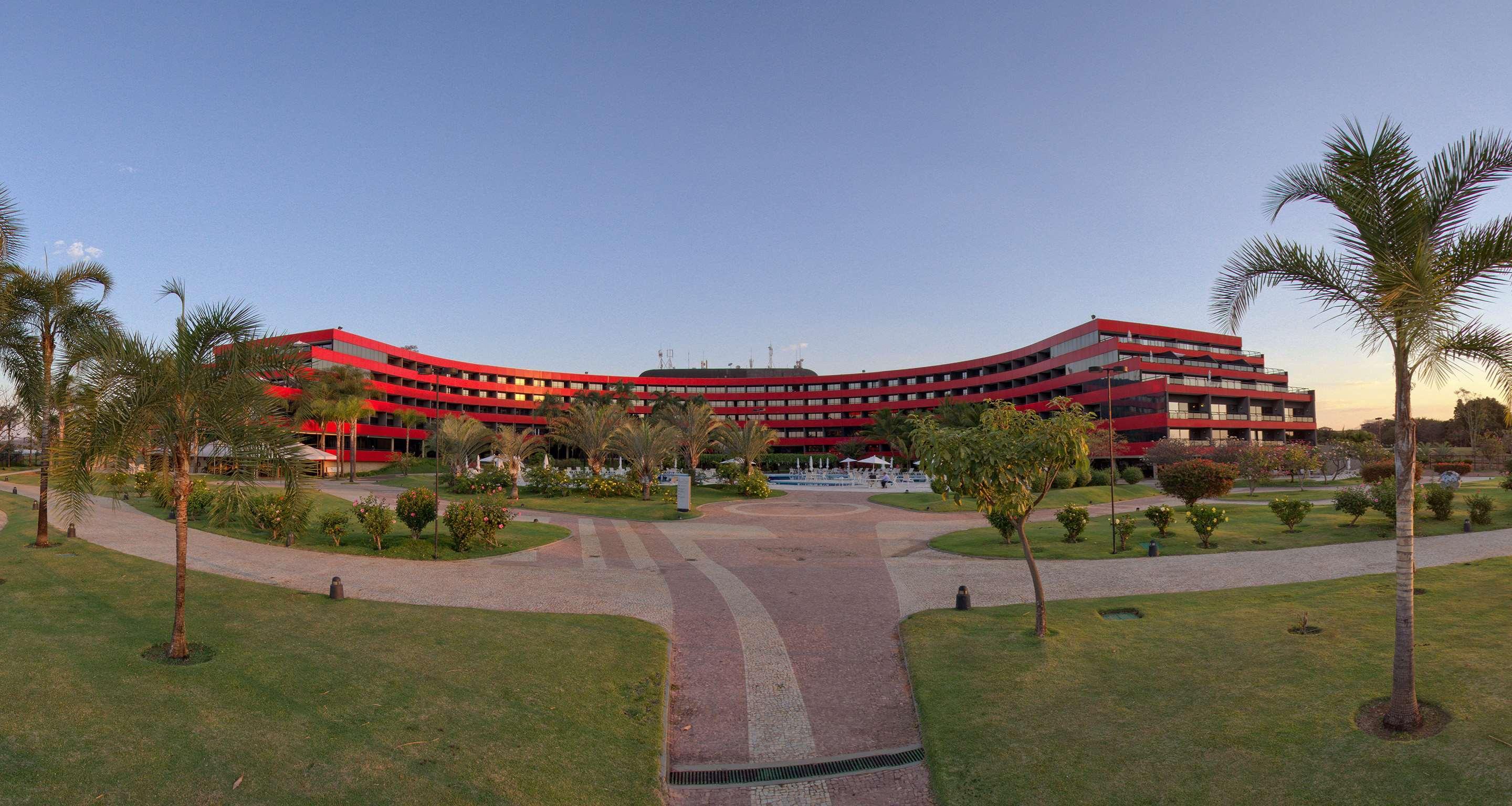 Royal Tulip Brasilia Alvorada Hotel Luaran gambar The photo shows a large, curved building with a vibrant red exterior. It appears to be a hotel or resort, surrounded by well-maintained gardens and green spaces. The landscape includes manicured lawns, small trees, and pathways, suggesting a pleasant