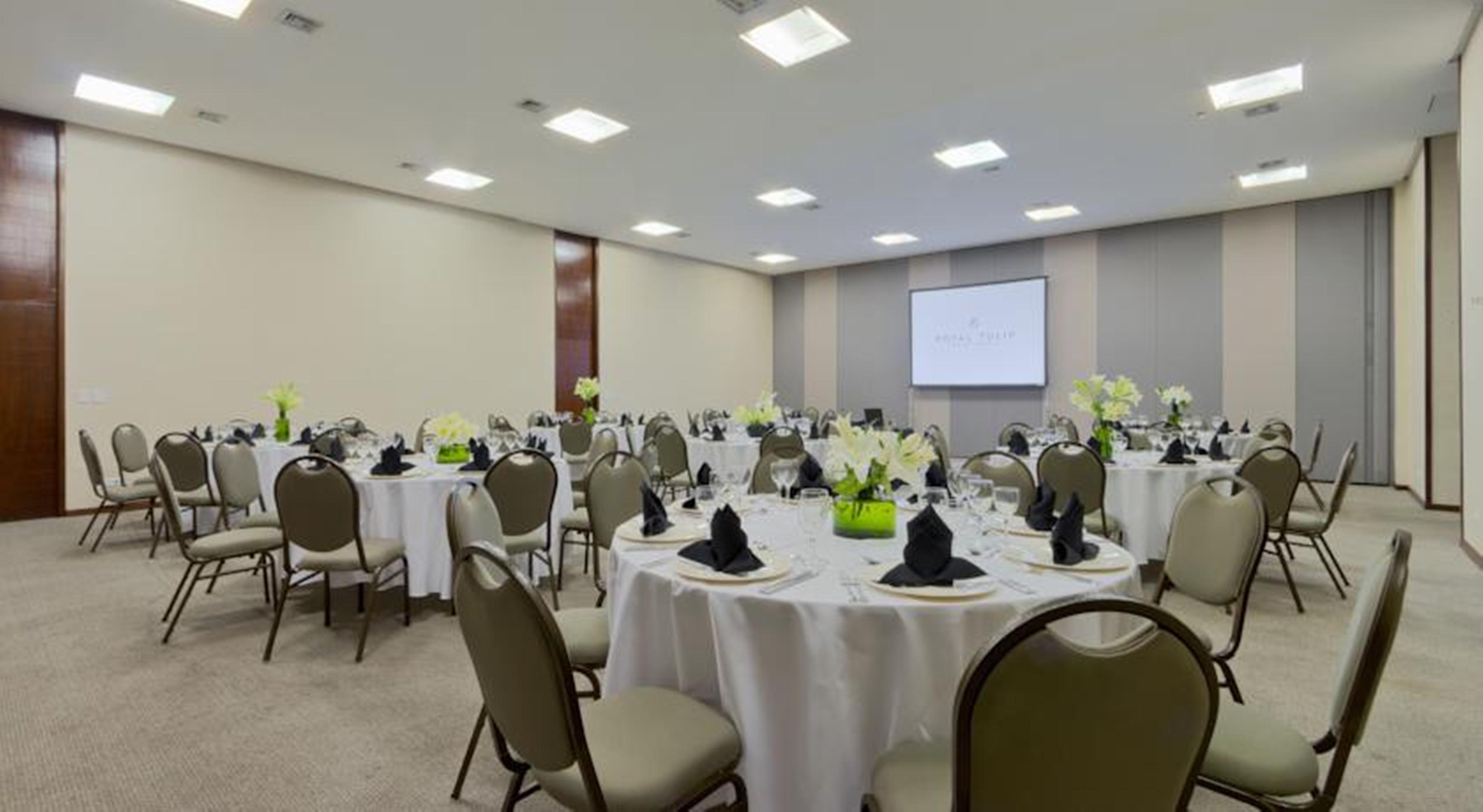 Royal Tulip Brasilia Alvorada Hotel Luaran gambar The photo shows a banquet or conference room set up with multiple round tables covered in white tablecloths. Each table is arranged with black napkins and centerpieces featuring flowers, likely in shades of green and white. The room has beige walls a