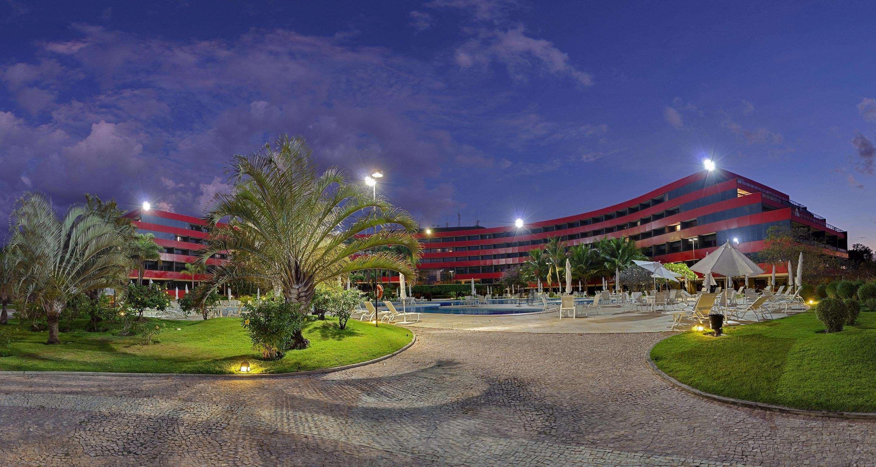 Royal Tulip Brasilia Alvorada Hotel Luaran gambar The photo shows a panoramic view of a hotel or resort complex, characterized by its modern architectural design with smooth, curved lines and a vibrant red color. It’s set in a landscaped area featuring palm trees and green grass. The surroundings in