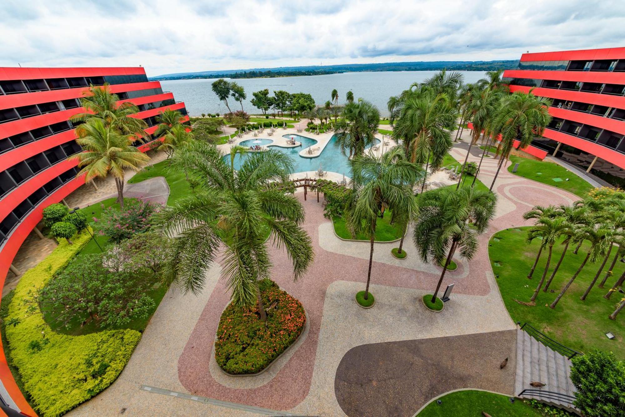 Royal Tulip Brasilia Alvorada Hotel Luaran gambar The photo shows a scenic view of a courtyard area in a residential complex. There are multiple red buildings surrounding a pool area, which is surrounded by palm trees and landscaped gardens. The pool features a curvy design, and there are pathways m