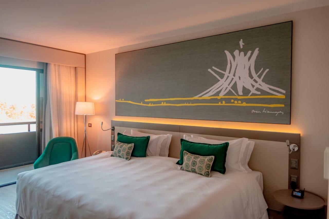 Royal Tulip Brasilia Alvorada Hotel Luaran gambar The photo shows a modern hotel room featuring a large bed with white bedding and two green accent pillows. Above the bed, there is a large piece of artwork depicting a stylized mountain or volcanic shape with yellow accents. The room has a contempora